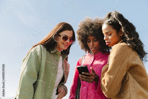 Fototapeta A group of three female friends standing and looking at a phone that one of them is holding while one of them is smiling