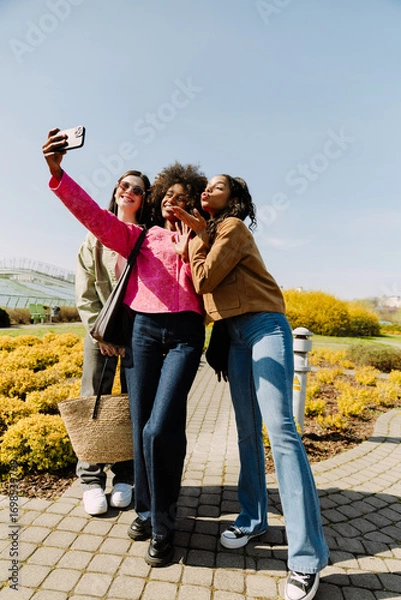 Fototapeta A group of three female friends smiling and posing while looking at a phone held by one of them