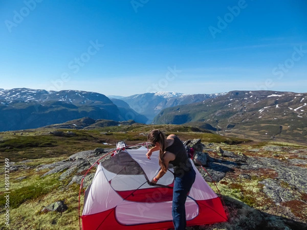 Obraz A woman assembles a red and white tent on a rocky mountaintop, preparing for a night of wild camping with a breathtaking view of a Norwegian fjord and distant, snow-capped mountains in the background.