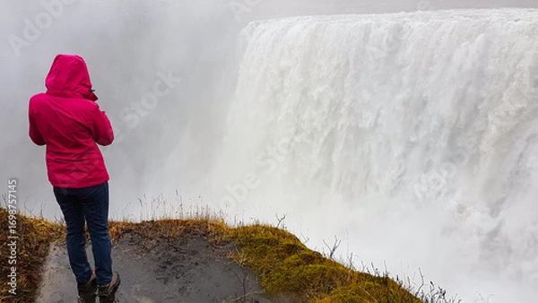Obraz A female tourist in a bright pink raincoat stands at the precipice, photographing the immense and powerful Dettifoss waterfall as it disappears into a thick cloud of mist in Iceland's wilderness.