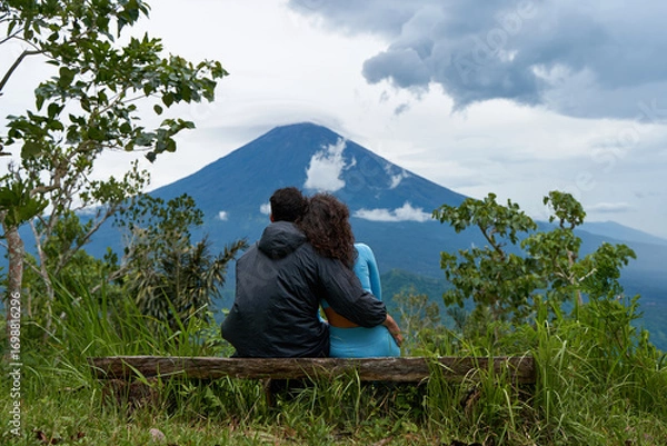 Fototapeta A couple of lovers are sitting on a bench, hugging each other and enjoying the view of the popular sacred cloud-covered Mount Agung on the island of Bali.