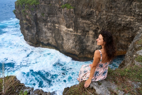 Fototapeta A beautiful woman in a pink dress sits on a cliff above the ocean on the island of Nusa Penida. Devil's Billabong an incredibly wonderful lagoon with splashes from the waves.