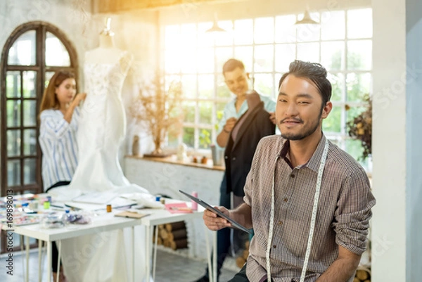 Obraz Young handsome asian confident fashion designer-clothes man with tablet pc and team are working at studio. Happy work for successful . Teamwork fashion designer concept