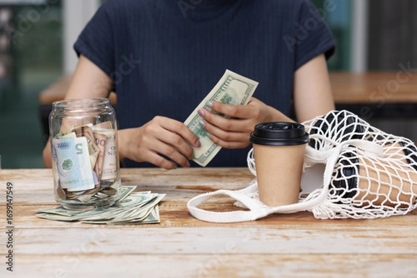 Fototapeta A person saves money by placing cash into a jar while sitting at a wooden table with a coffee cup and reusable net bag, symbolizing financial planning, savings, and eco-conscious living.
