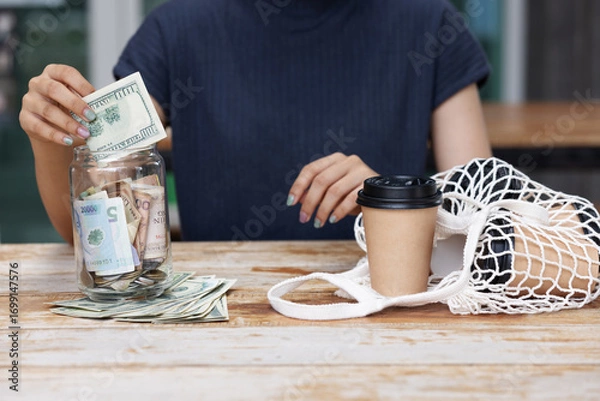 Fototapeta A person saves money by placing cash into a jar while sitting at a wooden table with a coffee cup and reusable net bag, symbolizing financial planning, savings, and eco-conscious living.