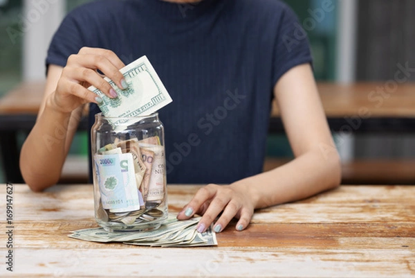 Fototapeta A person places dollar bills into a glass jar filled with cash on a wooden table, representing saving money, financial planning, and building future security through budgeting and smart spending.