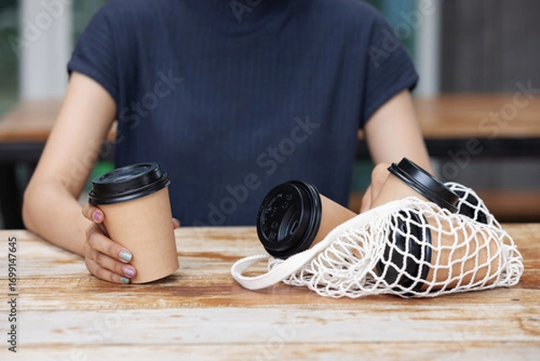 Fototapeta A person holds a takeaway coffee cup while more cups rest inside a reusable net bag on a wooden table, symbolizing eco-friendly habits, sustainability, and modern lifestyle choices.