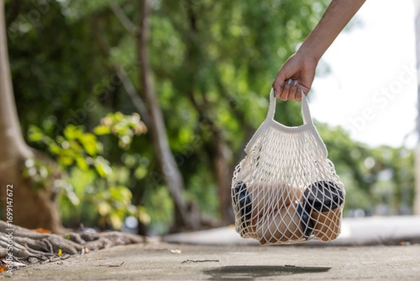 Fototapeta A hand carries a reusable net bag filled with takeaway coffee cups along a tree-lined street, symbolizing eco-friendly lifestyle choices, sustainability, and reducing single-use plastic waste.