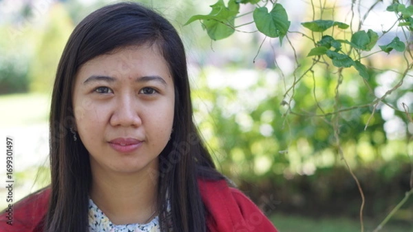 Fototapeta Portrait of a young Asian woman wearing a red jacket and floral blouse, standing outdoors with green plants and natural background, looking calm and confident