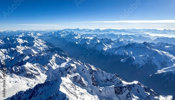 Fototapeta Snow-capped mountain range with valleys below, under a clear blue sky, some distant haze visible