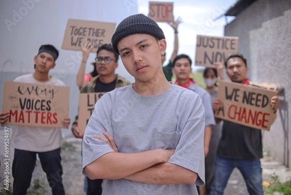 Fototapeta Young Asian Man With Black Beanie Crossed Arms Looking At Camera With Group Of Protestors