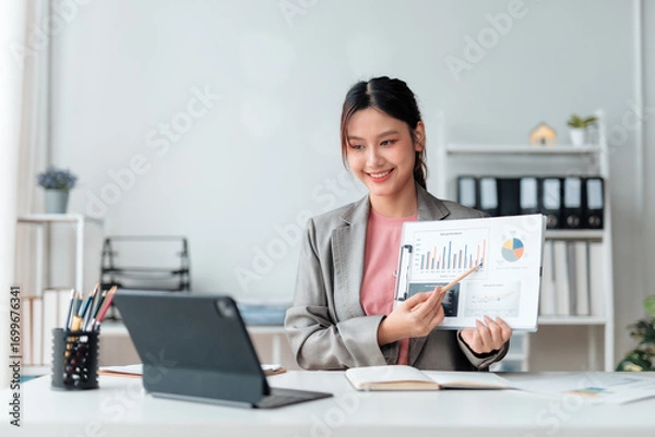 Fototapeta Young professional woman explaining financial charts and graphs during an online meeting using a digital tablet in her modern office