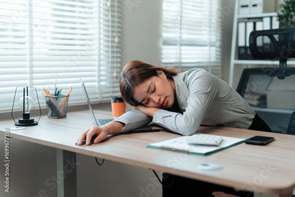 Fototapeta Young Asian office worker napping at her desk, finding a moment of rest after long hours of focused work on her laptop