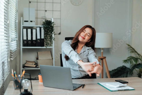Fototapeta Asian office worker relaxing after working with laptop, stretching arms at workplace, feeling happy and smiling