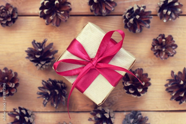 Obraz Christmas surprise/ gift wrapped in craft paper tied with a red bow against the background of fir cones