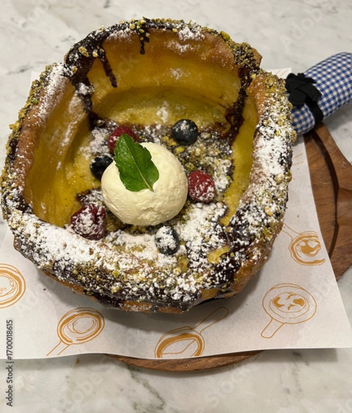 Fototapeta Delicious pancake with vanilla ice cream as dessert. Topped with fresh strawberry, crumble, chocolate sauce and icing powder on preheat Cast-iron skillet.