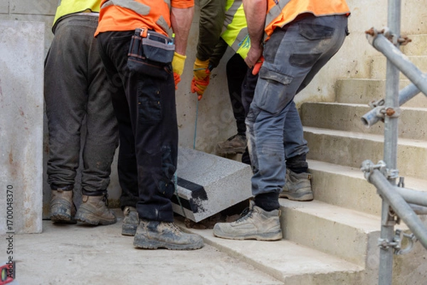 Fototapeta Team of builder workers lifting heavy concrete block for stairs construction. Teamwork and determination as a path to success concept.