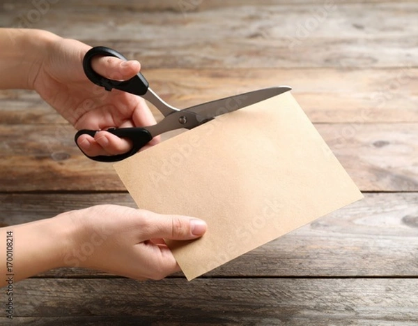Fototapeta Hands using black scissors to cut blank brown paper envelope on rustic wooden table surface