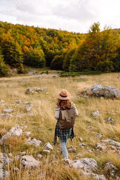 Obraz A rear view of a female hiker with a backpack and a hat walking along a rocky trail at sunset. Young woman enjoying the mountain scenery, admiring the views. Adventure, freedom and weekend concept.