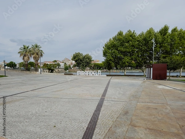 Fototapeta The open square by the Tua River in Mirandela, Portugal, is framed by palm trees and city landmarks. Inviting for gatherings, this riverside space connects people to nature and urban charm.