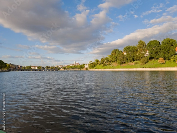 Fototapeta Wide view of the Tua River in Mirandela, Portugal, with the pedestrian bridge and city skyline in the distance. Evening clouds and tranquil water highlight this urban landscape and natural beauty.