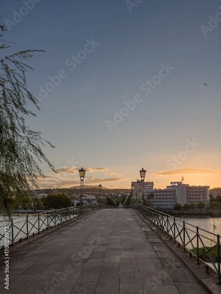 Fototapeta Pedestrian bridge over the Tua River in Mirandela, Portugal, leads to the city center at sunset. Evening light, calm water, and urban skyline create a tranquil and scenic connection.