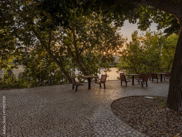 Fototapeta In Mirandela, Portugal, the riverside square by the Tua features outdoor tables and chairs for relaxation, framed by scenic bridges, gardens, and the charm of this northern town.