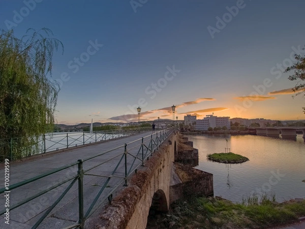 Fototapeta Pedestrian bridge over the Tua River in Mirandela, Portugal, leads to the city center at sunset. Evening light, calm water, and urban skyline create a tranquil and scenic connection.