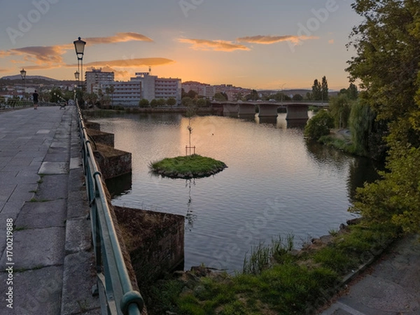 Fototapeta Pedestrian bridge over the Tua River in Mirandela, Portugal, leads to the city center at sunset. Evening light, calm water, and urban skyline create a tranquil and scenic connection.