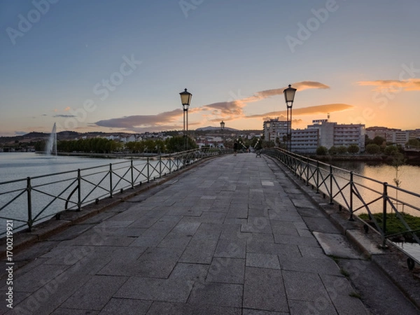 Fototapeta Pedestrian bridge over the Tua River in Mirandela, Portugal, leads to the city center at sunset. Evening light, calm water, and urban skyline create a tranquil and scenic connection.