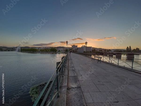 Fototapeta Pedestrian bridge over the Tua River in Mirandela, Portugal, leads to the city center at sunset. Evening light, calm water, and urban skyline create a tranquil and scenic connection.
