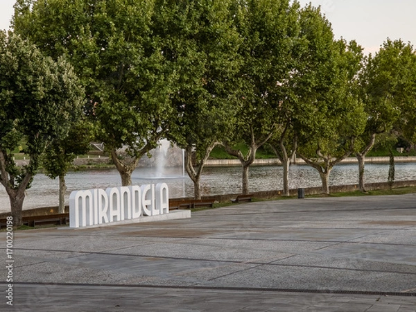 Fototapeta Mirandela, Portugal: The riverside square with large trees and the Tua River creates a welcoming urban space, where the iconic city sign invites residents and visitors to enjoy leisure and nature.
