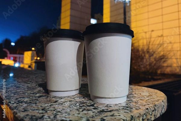 Obraz Two blank coffee cups resting on a stone surface at night in an urban area with city lights in the background