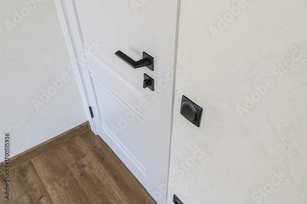 Fototapeta Close-up of a white door with a sleek black handle, paired with a black switch against a textured cream wall. Wooden floor trim visible