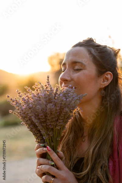 Fototapeta Woman smelling and enjoying lavender bouquet at sunset in field, vertical capture