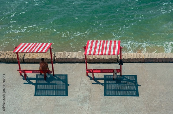 Obraz Man Sitting Under Red Striped Shade by the Sea
