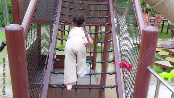 Obraz Happy Young Asian child girl climbing a rope bridge at a playground, showcasing outdoor play and adventure.