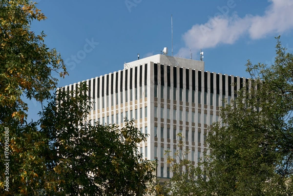 Fototapeta Upward view of a modern office tower with vertical black stripes on the roofline, framed by green trees, Saskatoon, Canada.