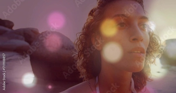 Fototapeta Wading woman wearing white swimsuit and necklace at beach shoreline by rocks with lens flare bokeh