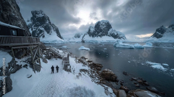 Fototapeta Couple in winter gear walking hand in hand on snowy deck by icy coastal water with towering iceberg and jagged mountain peaks under overcast sky, serene cold mood