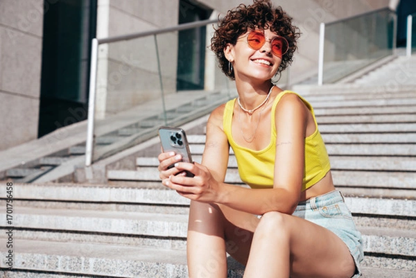 Obraz Young beautiful smiling hipster woman in trendy summer clothes. Carefree woman with curls hairstyle, in the street. Positive model holds mobile phone, looks at cellphone screen, uses smartphone apps