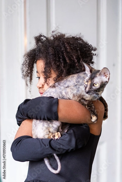 Fototapeta Playful Afro-American girl with curly afro hair embraces her pet cat, showcasing a heartwarming bond in a bright indoor setting with soft textures and warm ambiance