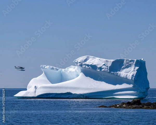 Fototapeta A small plane passes near a large iceberg that is close to shore.