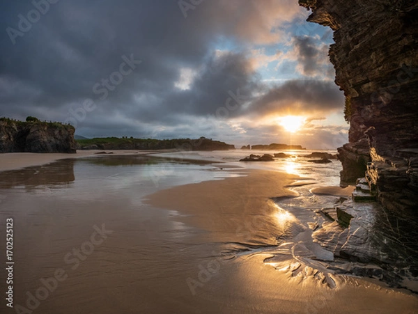 Fototapeta Dramatic cliffs and sandy shore of Playa de las Catedrales in Galicia, Spain, at sunset with golden light breaking through clouds, capturing natural beauty, solitude, and wonder