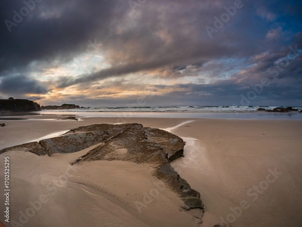 Fototapeta Dramatic cliffs and sandy shore of Playa de las Catedrales in Galicia, Spain, at sunset with golden light breaking through clouds, capturing natural beauty, solitude, and wonder