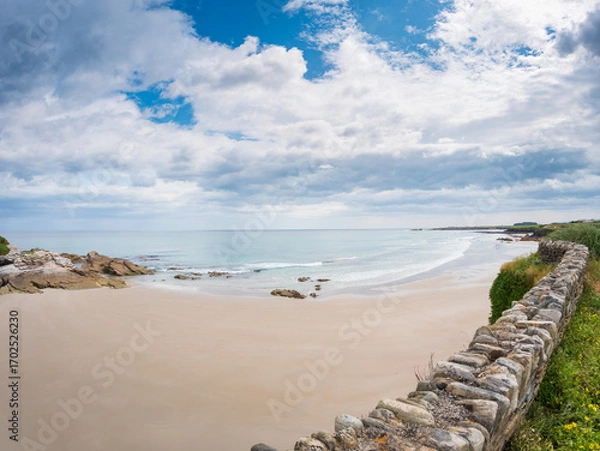 Fototapeta Wide view of Playa de las Catedrales in Galicia, Spain, showing sandy beach, rocky formations, calm waves, stone wall, and dramatic cloudy sky along the Atlantic coast.