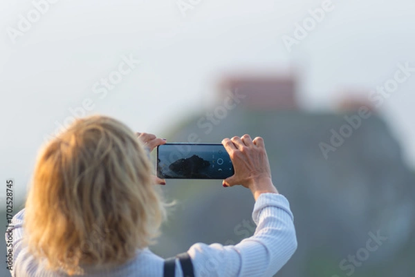 Fototapeta Tourist taking a smartphone photo of San Juan de Gaztelugatxe in Basque Country, Spain, with blurred rocky island and chapel in the background captured on a hazy day
