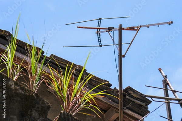 Fototapeta An old, rusty television antenna stands tall against a clear blue sky, overlooking the rooftops of Xian Guang Military Kindred Village Cultural Park in Taiwan. A nostalgic glimpse of the past.