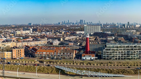Fototapeta A coastal cityscape of Scheveningen, with a prominent red lighthouse in the foreground. Modern buildings line the horizon of The Hague under a clear blue sky.