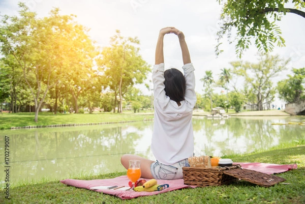 Fototapeta Closeup of young woman streching and relaxing with fruit basket and glass of orange juice in the park at weekend. Wellness. Health and food concept.